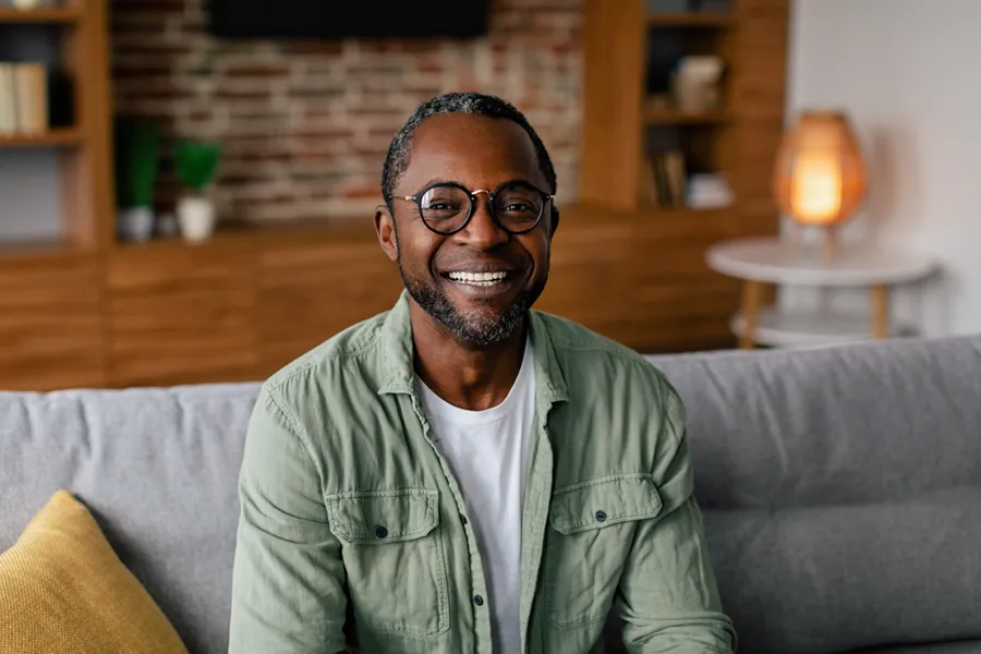 Andropause-Doctor A middle aged black man in a green button up shirt sits on his couch smiling, happy with his treatment for andropause and low testosterone from Healthy Aging Medical Centers of New Jersey in Northern New Jersey.
