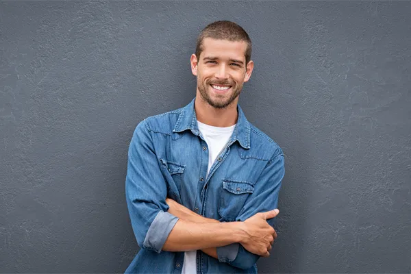 Hormones-for-Men-Doctor A man in a denim shirt stands smiling against a gray-blue wall, pleased with his testosterone hormone therapy from Healthy Aging Medical Centers of New Jersey in Northern New Jersey.