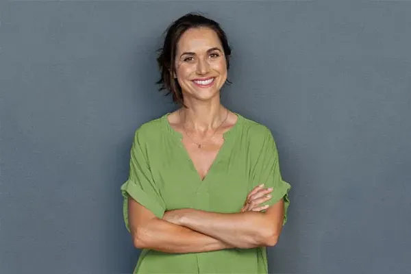 Hormones-for-Women-Doctor A woman in a green shirt stands smiling against a gray-blue wall, pleased with her hormone therapy from Healthy Aging Medical Centers of New Jersey in Northern New Jersey.
