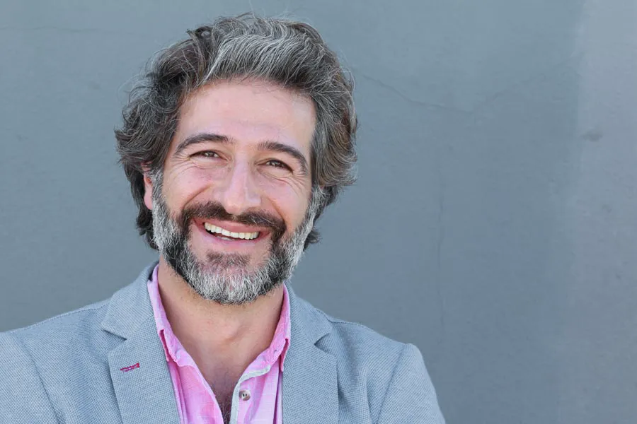 Hypogonadism-Clinic A smiling man with salt and pepper hair and beard wearing a pink shirt and gray blazer stands agains a gray wall. Get hypogonadism treatment from Healthy Aging Medical Centers of New Jersey in Northern New Jersey.