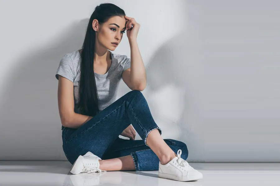 PCOS-Doctor A young brunette woman in a gray t-shirt and jeans sits against a gray wall with her head resting on her hand and a distressed expression. Get help for Polycystic Ovarian Syndrome (PCOS) from Healthy Aging Medical Centers of New Jersey in Northern New Jersey.