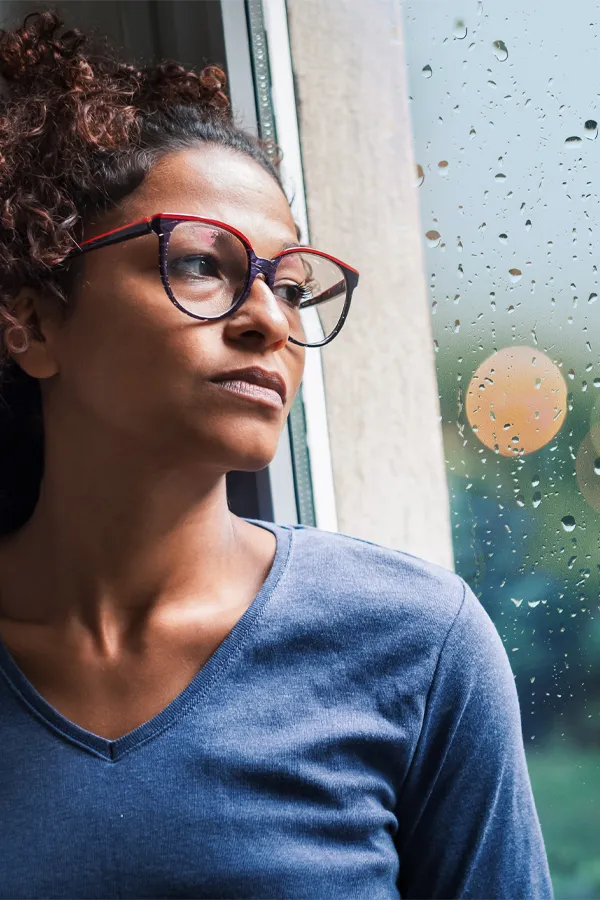 PMDD-Treatment A black woman with with a somber expression and glasses and a gray shirt stares out the window on a rainy day. Get treatment for Premenstrual Dysphoric Disorder from Healthy Aging Medical Centers of New Jersey in Northern New Jersey.