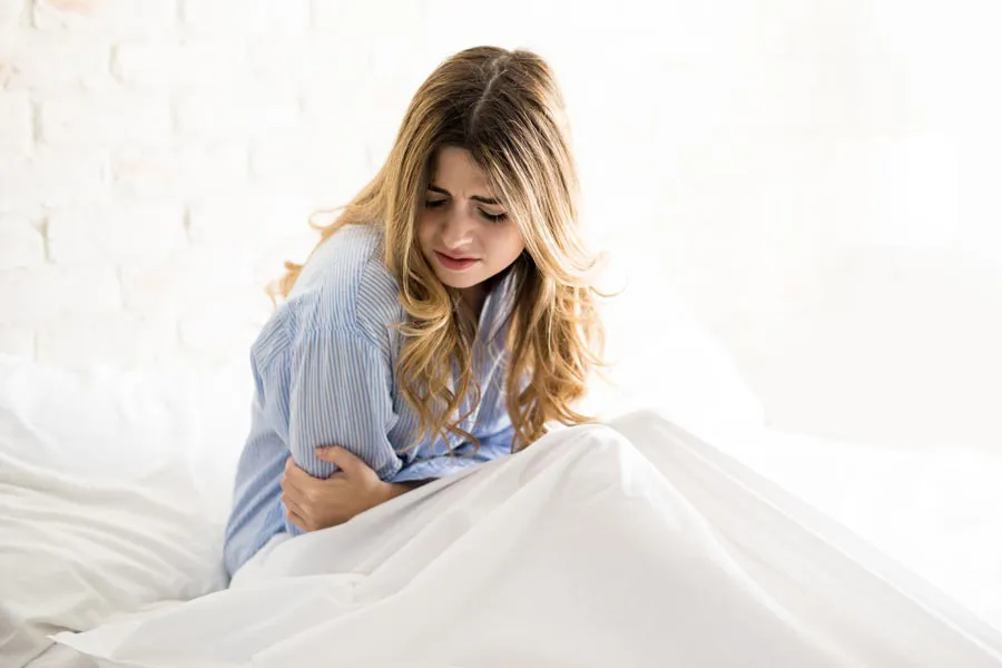 PMS-Therapy A woman with long blonde hair sits in her bed holding her stomach in pain, considering getting care for PMS from Healthy Aging Medical Centers of New Jersey in Northern New Jersey.
