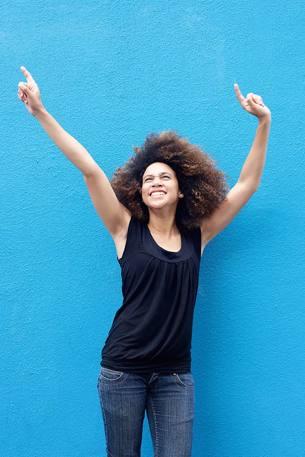PMS-Treatment A woman in a dark blue tank top standing in front of a bright blue wall, raising her arms in celebration of relief from PMS from Healthy Aging Medical Centers of New Jersey in Northern New Jersey.