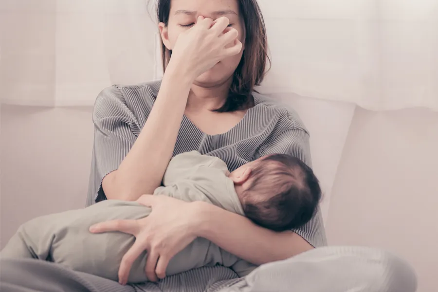 Postpartum-Hormone-Imbalance-Clinic A woman nursing an infant on couch rubs her eyes with exhaustion. Get treatment for postpartum hormone imbalance from Healthy Aging Medical Centers of New Jersey in Northern New Jersey.